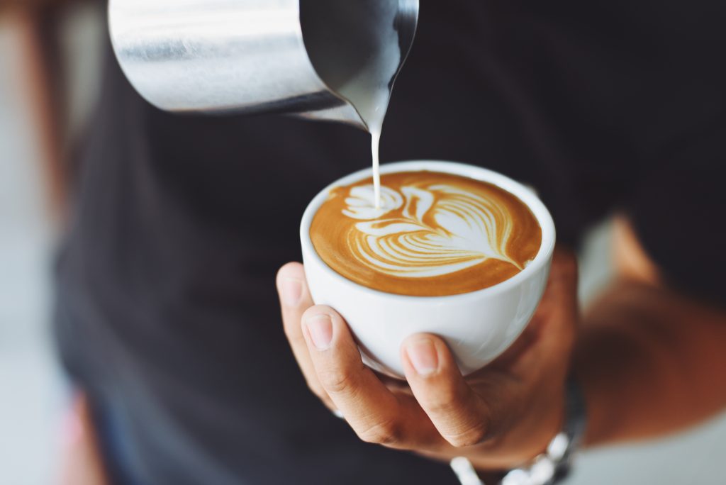 close up shot of a coffee shop worker creating a floral design with cream in a small cup of coffee.