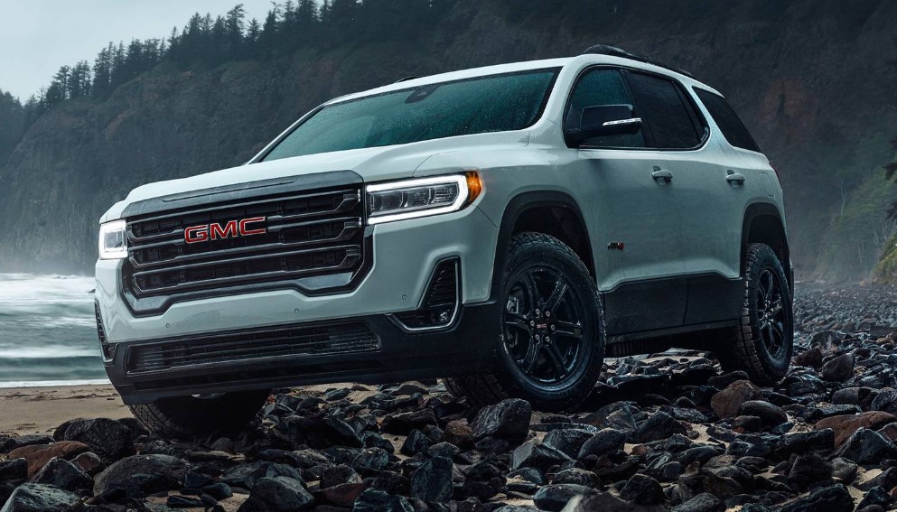 Brand new white 2023 GMC Acadia driving over a rocky beach with lushes green forest in the background.