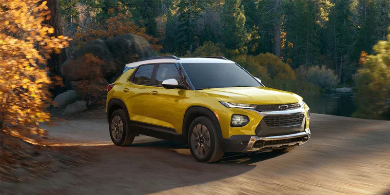 Pictured is a brand new yellow 2023 Chevrolet Trailblazer driving down a dirt road with trees all around.