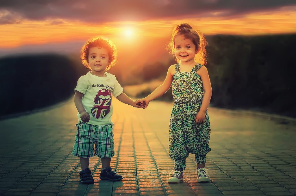 two little kids, a girl and a boy, holding hands smiling at the camera with a sunset in the background.