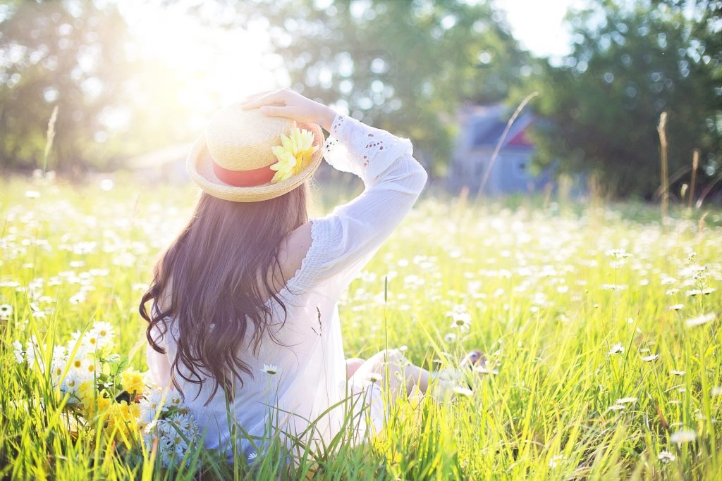 Woman, with her back facing you, is sitting in an open field full of wildflowers with the sun shinning on her. 