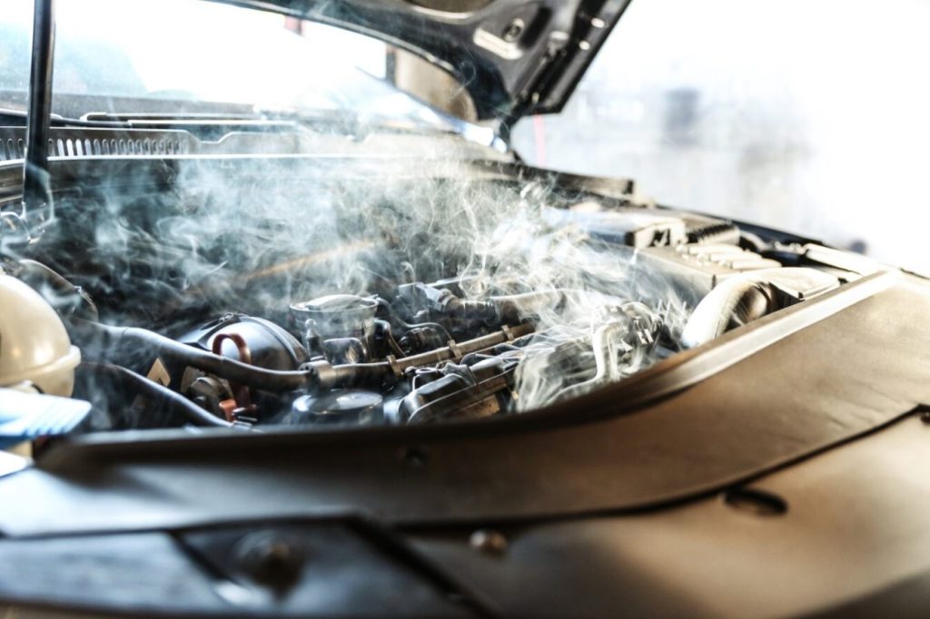 The image features a close-up view of a car's engine, with steam or smoke billowing from it. The engine appears to be hot or in the process of cooling down. The scene may suggest that the car has been recently turned off or has been running for a while.