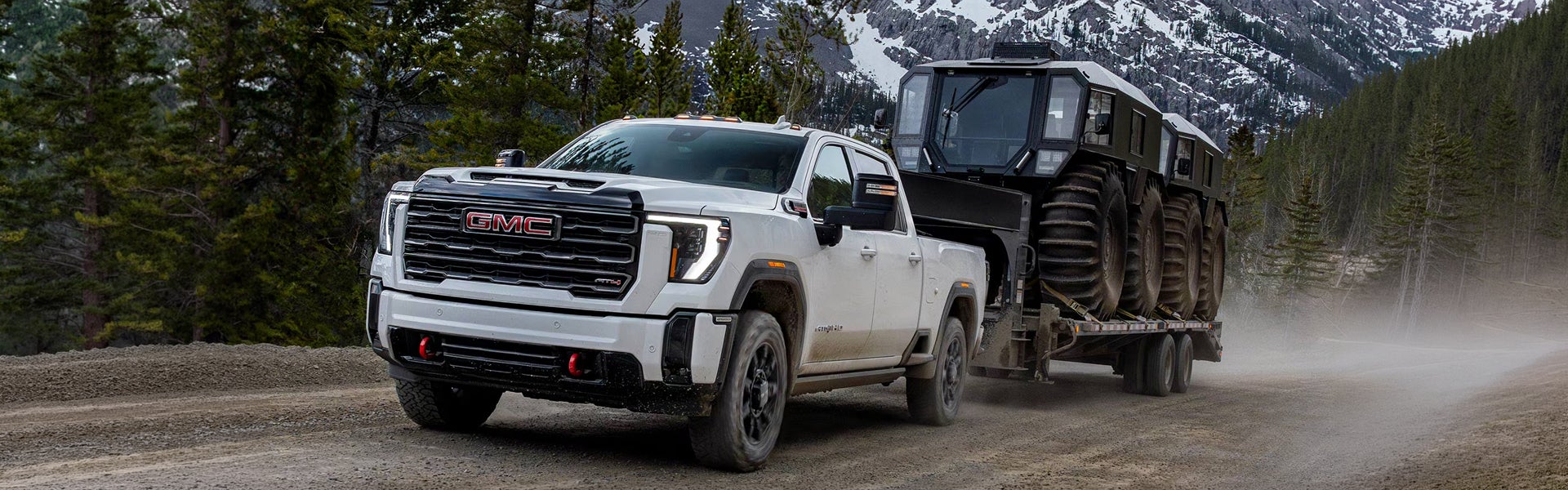 White 2026 GMC Sierra 3500HD towing a heavy tracked trailer through snowy mountains