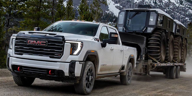 White 2026 GMC Sierra 3500HD towing a tracked trailer through snowy mountains