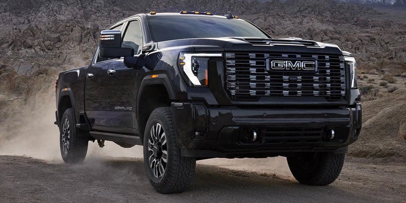 Black 2026 GMC Sierra 3500HD driving on a dusty gravel road, front three-quarter view