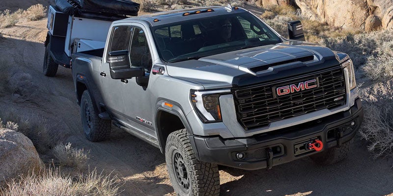Silver 2026 GMC Sierra 3500HD towing a trailer with a tracked vehicle over rocky terrain
