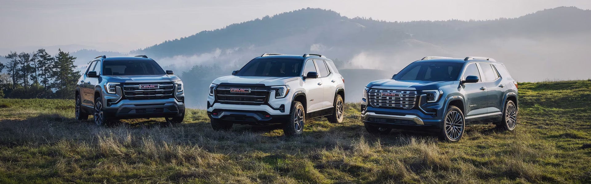 The 2027 GMC Terrain lineup parked on open grassland below misty hills, front three-quarter view