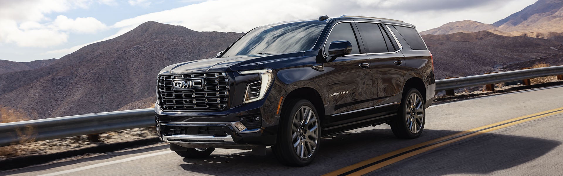 2027 GMC Yukon Denali driving on a mountain road, dark exterior, front three-quarter view